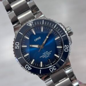 Close-up of a polished stainless steel Oris diver’s watch with a vivid blue dial, luminous hands and markers, and a numbered rotating bezel against a soft gray background.