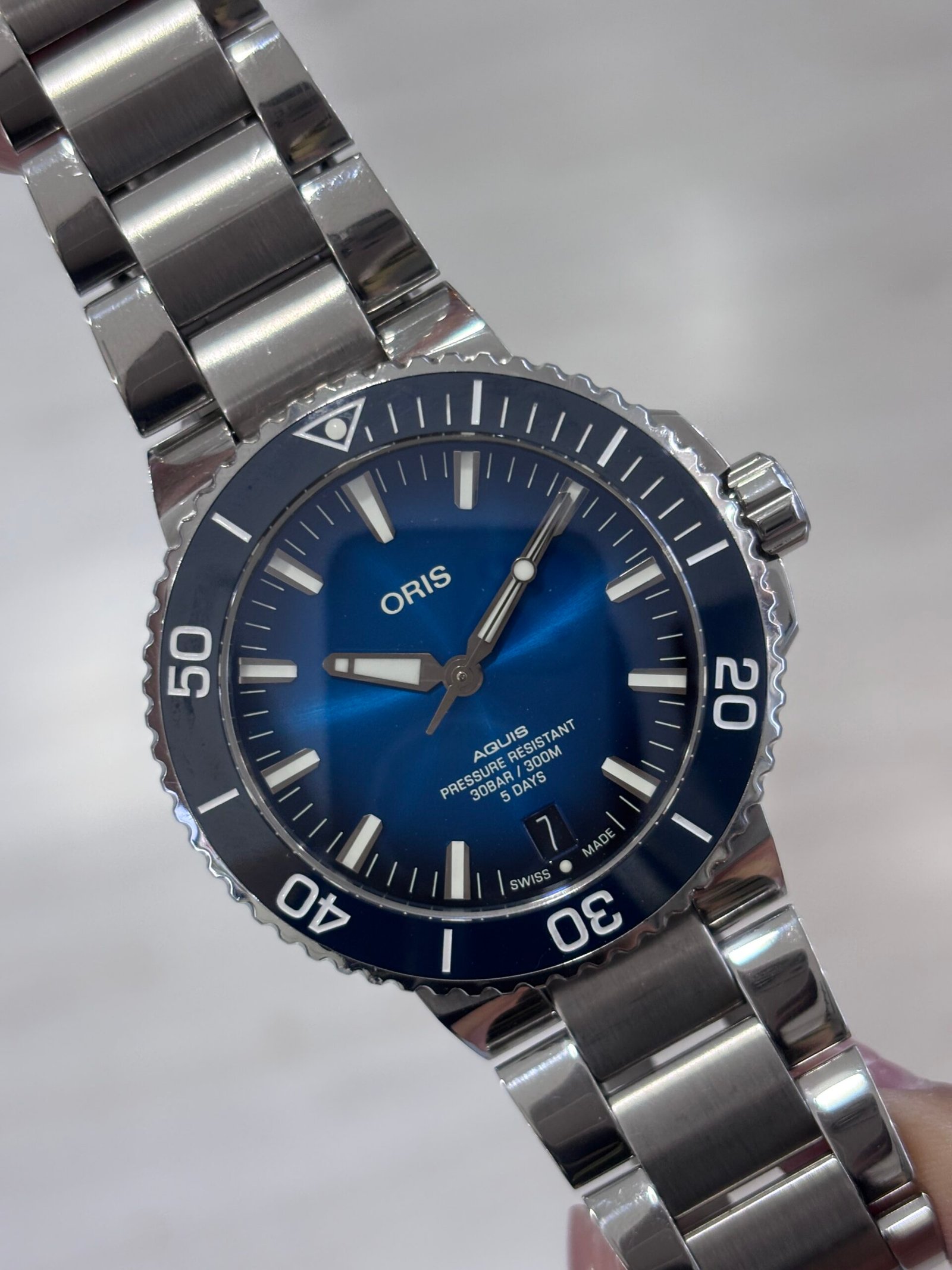 Close-up of a polished stainless steel Oris diver’s watch with a vivid blue dial, luminous hands and markers, and a numbered rotating bezel against a soft gray background.