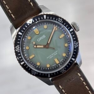 Close-up of an Oris diver’s watch with a muted green dial, gold-tone markers, black numbered bezel, and stitched brown leather strap, conveying rugged elegance.