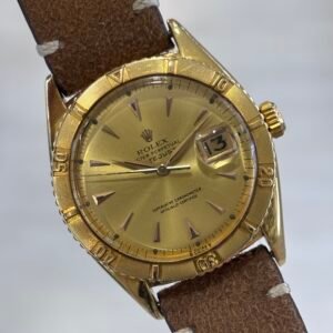 Vintage gold Rolex wristwatch with a textured bezel, cream dial, date magnifier, and worn brown leather strap, photographed against a pale background.