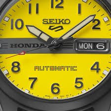 Close-up of a bright yellow Seiko Honda wristwatch dial with bold black numerals, silver hands, day-date window, and sporty racing details.