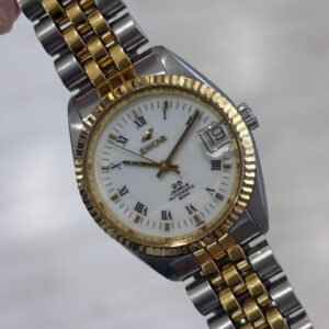 Close-up of a two-tone gold and silver Bulova wristwatch with Roman numerals, a fluted bezel, and date window, held against a soft gray background. Its polished finish feels classic and refined.