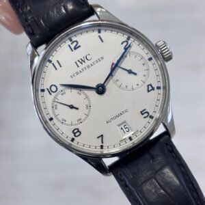Close-up of an IWC Schaffhausen automatic wristwatch with a silver dial, blue hands, date display, and black leather strap, held against a soft gray background.