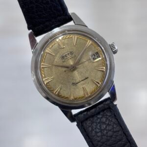 A vintage wristwatch with a textured gold dial, slim hour markers, date window, silver case, and black leather strap, photographed against a pale background.