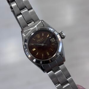 A hand holds a stainless-steel Rolex watch with a dark brown dial, gold hour markers, and date window, angled against a soft gray background.A hand holds a stainless-steel Rolex watch with a dark brown dial, gold hour markers, and date window, angled against a soft gray background.