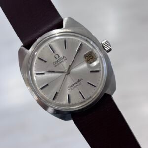 A hand holds a vintage silver Omega Seamaster Cosmic watch with a cream dial, date window showing 28, and dark brown leather strap.