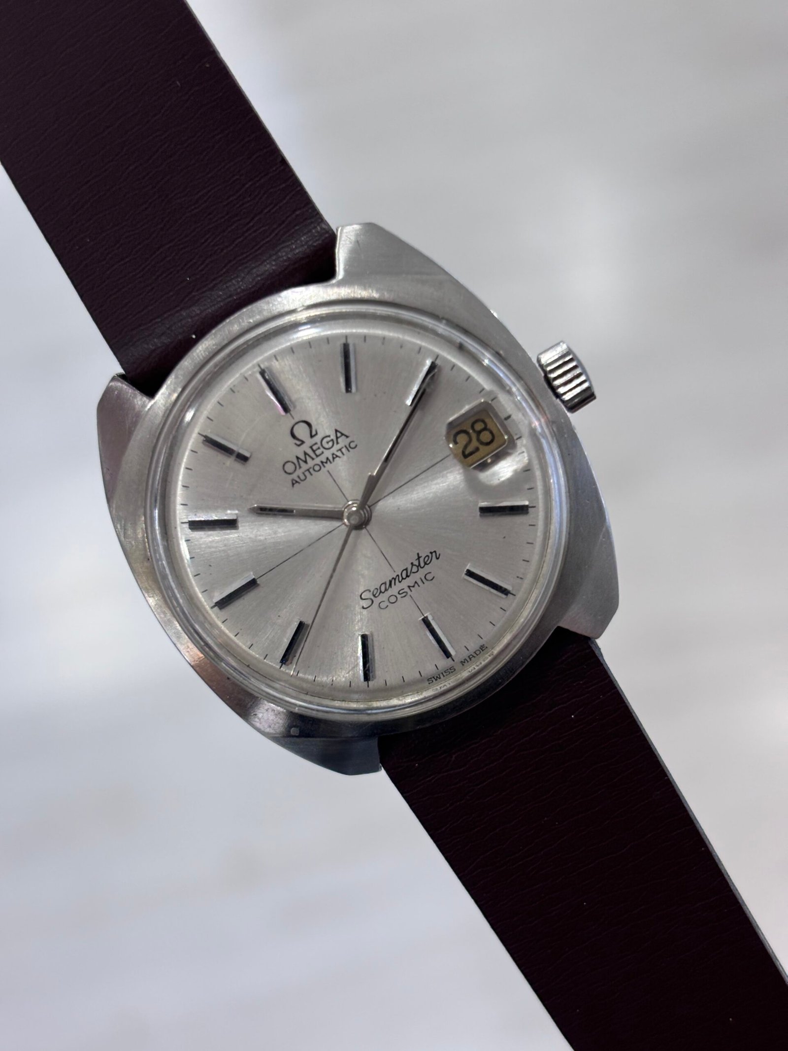 A hand holds a vintage silver Omega Seamaster Cosmic watch with a cream dial, date window showing 28, and dark brown leather strap.