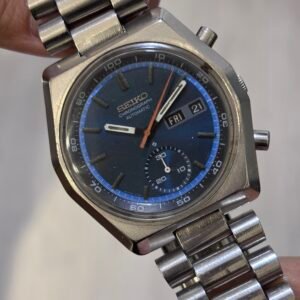 Close-up of a hand holding a vintage Seiko automatic wristwatch with a blue dial, day-date display, and stainless-steel bracelet; the worn finish adds a nostalgic tone.