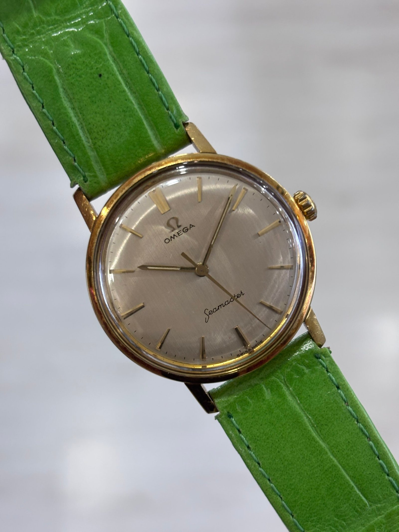 A vintage gold-tone Omega Seamaster wristwatch with a silver dial and bright green leather strap, photographed against a soft gray background.
