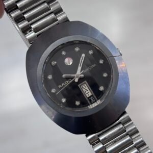 Close-up of a vintage Rado stainless-steel wristwatch with a dark jeweled dial and day-date window, angled against a soft gray background.