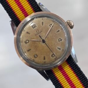 A vintage Omega wristwatch with a weathered cream dial, rose-gold-toned bezel, and black, red, and yellow striped fabric strap against a pale background.